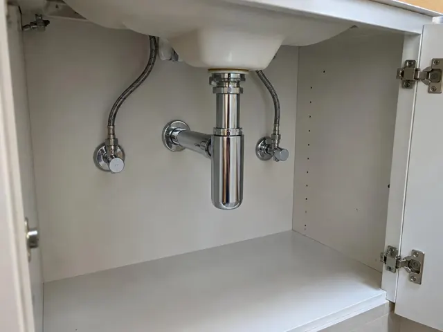 Neat under-sink cabinet with chrome drain pipes, flexible water lines and dry white panels in Haberfield